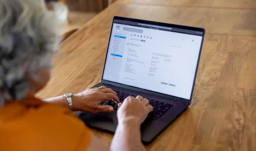 Vista por cima do ombro de uma mulher com cabelos grisalhos usando um notebook moderno sobre uma mesa de madeira.
