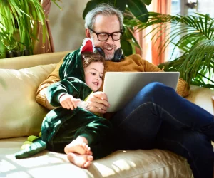 Homem faz uso de computador do lado de filha ou neta pequena, que sorri para o que ele está vendo