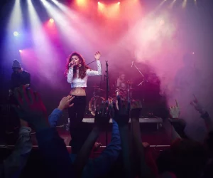 Uma cantora com cabelos cacheados no palco sob luzes coloridas e fumaça, com o público ao fundo com as mãos levantadas.