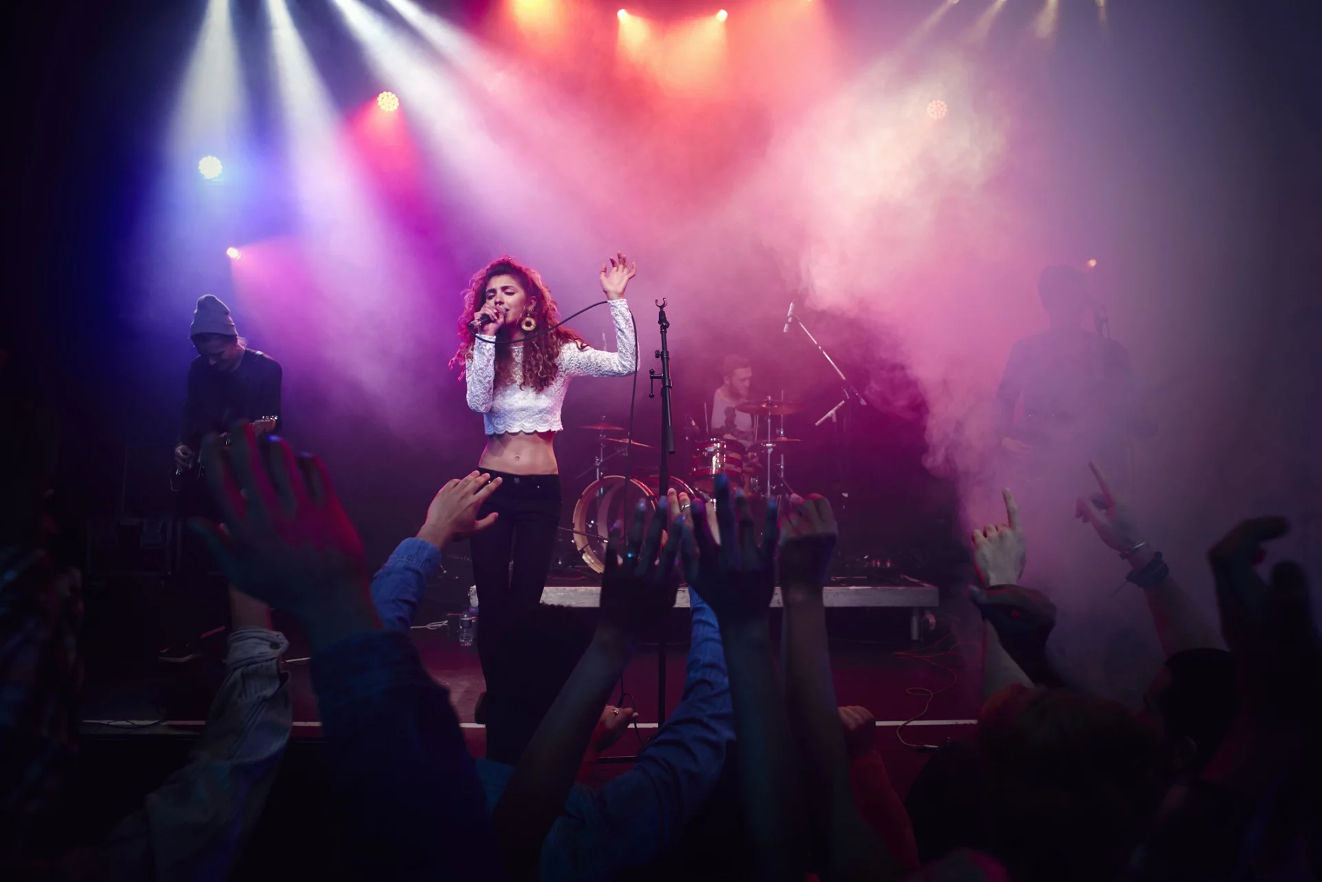 Uma cantora com cabelos cacheados no palco sob luzes coloridas e fumaça, com o público ao fundo com as mãos levantadas.