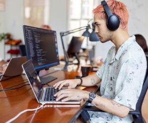 Jovem mexe com dois computadores enquanto ouve música no headphone porque a computação é uma das profissões em alta