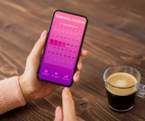 Pessoa segurando smartphone com aplicativo de calendário menstrual aberto, ao lado de uma xícara de café.