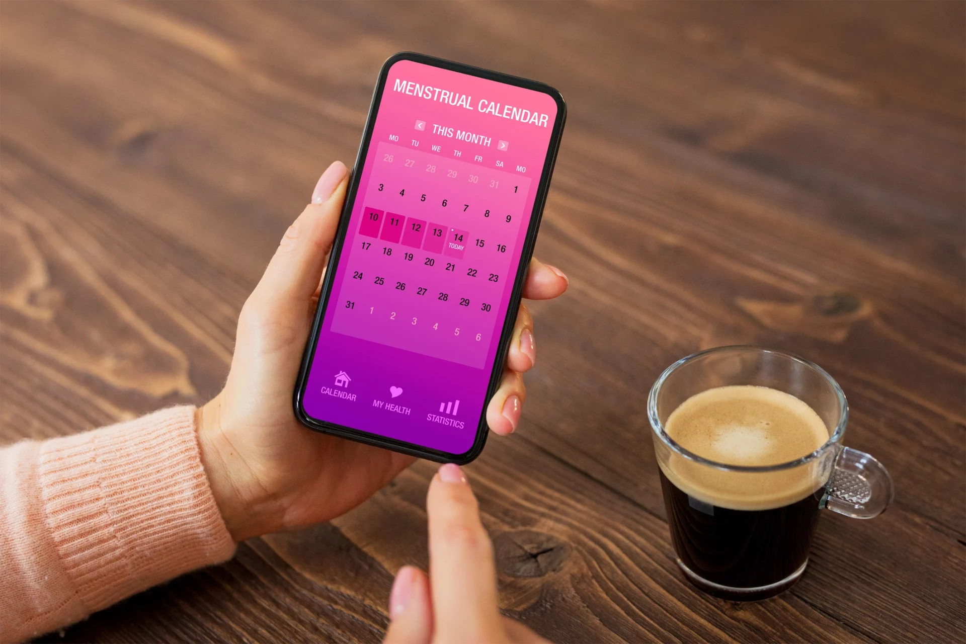 Pessoa segurando smartphone com aplicativo de calendário menstrual aberto, ao lado de uma xícara de café.