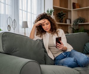 Mulher de cabelos cacheados sentada em um sofá verde, segurando um celular com uma mão e a outra na testa, demonstrando preocupação.