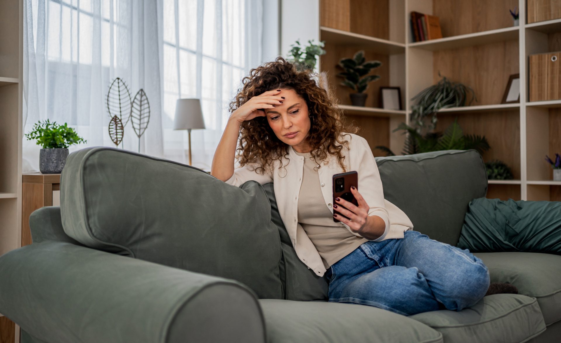 Mulher de cabelos cacheados sentada em um sofá verde, segurando um celular com uma mão e a outra na testa, demonstrando preocupação.
