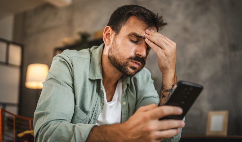 Homem jovem com barba e camisa verde, sentado, com expressão de cansaço e a mão na testa enquanto olha para a tela do celular.