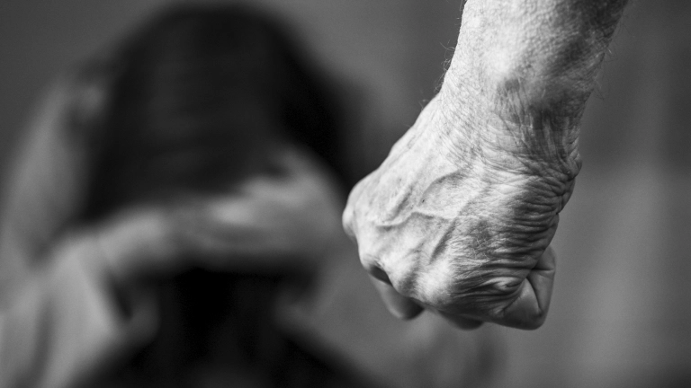 Mão de homem cerrada em punho no primeiro plano está fechada para agredir vítima feminina que se encolhe ao fundo. Essa foto em preto e branco simboliza o machismo e a violência contra a mulher