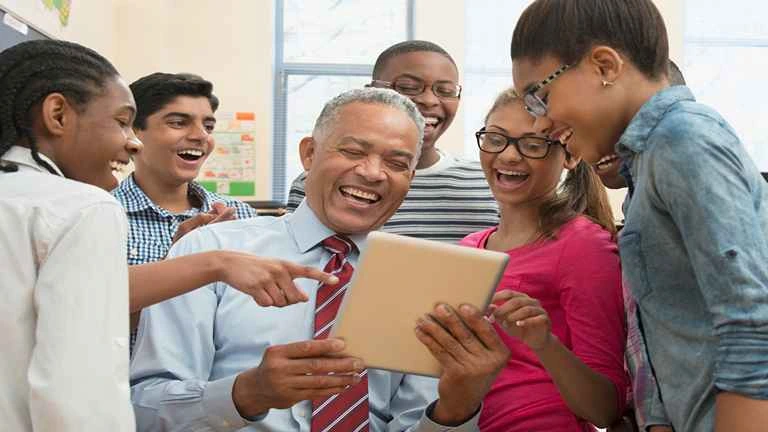 Homem segura um tablet e ri, está rodeado de jovens que fazem voluntariado on line e que querem auxiliá-lo com a tecnologia 