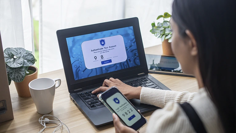 Mulher com celular em uma mão faz processo de autenticação para entrar no computador. Um dos princípios da segurança da informação inclui o cuidado com senhas e códigos