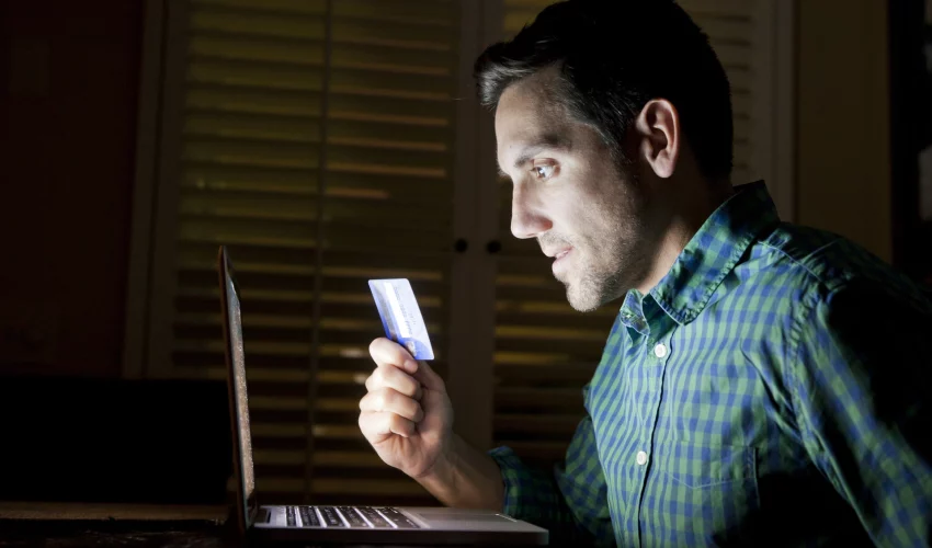 Homem olha de forma obcecada para o computador com o seu cartão na mão. Imagem pode representar o risco das apostas online