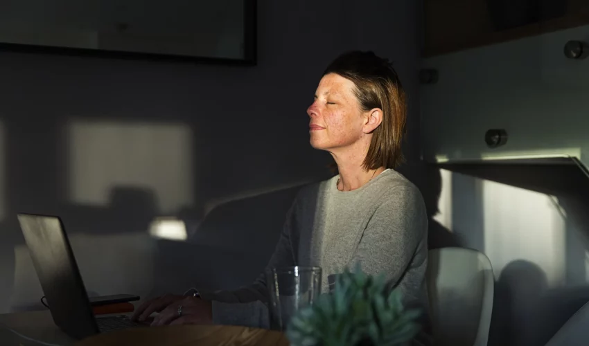 Mulher fecha os olhos na frente do computador e dá um sorriso leve enquanto o sol ilumina o seu rosto. Esta imagem representa um tempo de pausa neste janeiro branco para priorizar a sua saúde mental 