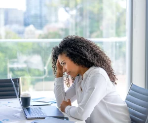 Mulher sentada em escritório na frente do computador com uma mão no rosto que mostra preocupação e cansaço. Esse é um dos sintomas do brain rot, palavra do ano