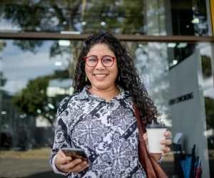 mulher sorri enquanto segura um celular e segura um café. A postura faz parecer que ela está fora de casa, no seu trabalho