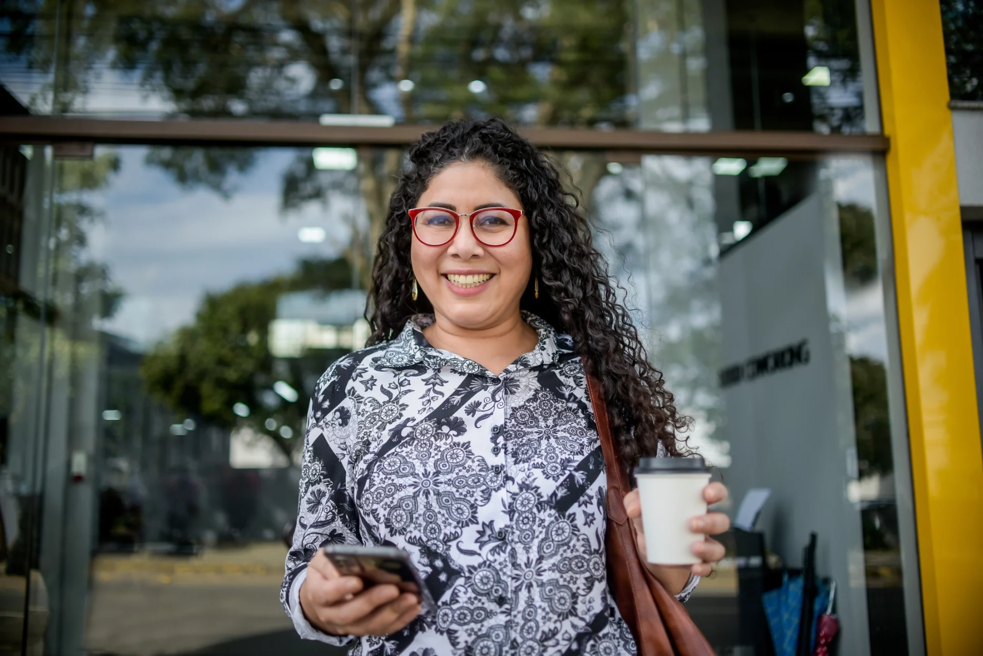 mulher sorri enquanto segura um celular e segura um café. A postura faz parecer que ela está fora de casa, no seu trabalho