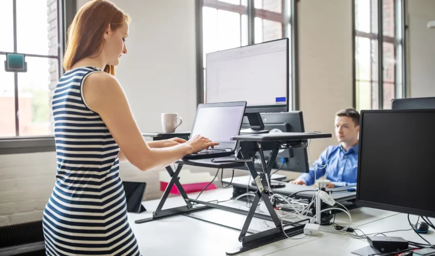 Mulher trabalha de pé em computador na mesa com apoio para ficar na altura dos olhos dela. É uma forma de ter ergonomia no trabalho