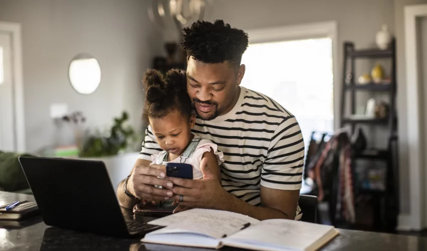 Homem mexe em seu celular com a filha no colo, para auxiliá-la no uso da tecnologia durante a infância