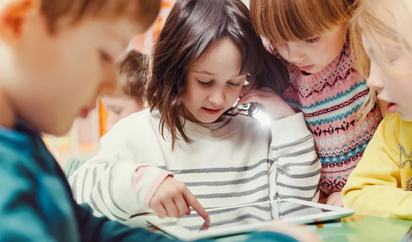 Criança pequena faz uso de tablet ao lado de seus colegas. O uso da tecnologia na infância auxilia na leitura de histórias