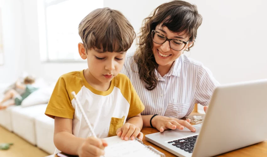 Mulher para filho que está fazendo lição ao seu lado. Ela tem um computador em sua frente, para auxiliar o uso da tecnologia na infância