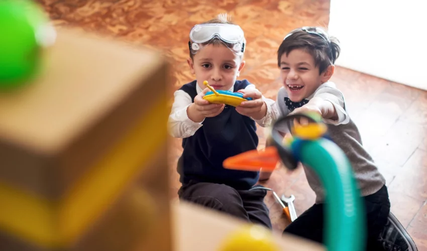 Duas crianças sentadas no chão apontam um controle para uma caixa, sorrindo. Indicativo de que estão brincando e se divertindo