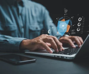 Mãos digitando em laptop com holograma de cérebro e gráficos rotulados "AI" acima do teclado.