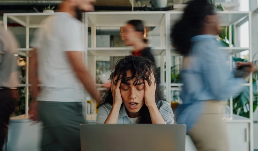 Uma mulher com as mãos no rosto e expressão de desespero em frente a um laptop, enquanto vultos de pessoas se movendo rapidamente passam ao seu redor.