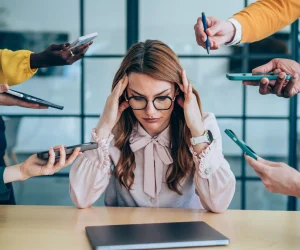Uma mulher sentada à mesa com as mãos na cabeça em sinal de estresse, cercada por várias mãos segurando smartphones e tablets em sua direção.