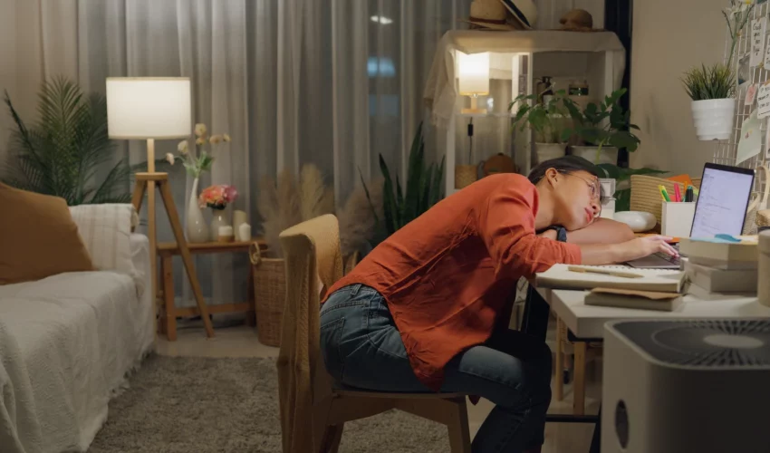 Uma mulher jovem com óculos dormindo debruçada sobre o teclado de um laptop em uma mesa de trabalho em casa durante a noite.