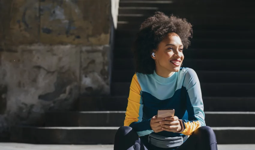 Uma mulher jovem com cabelo afro sentada ao ar livre em degraus de pedra, sorrindo e olhando para o lado enquanto segura um celular de forma relaxada.