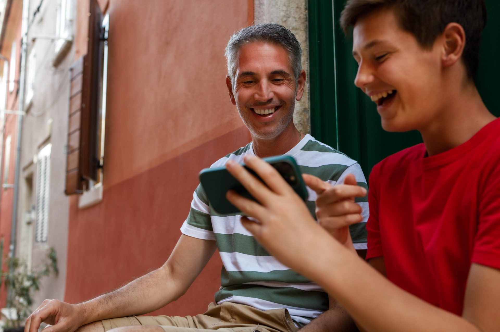 Pai e filho adolescente sentados na rua, rindo e compartilhando experiências para fazer com filhos através de um vídeo no celular.