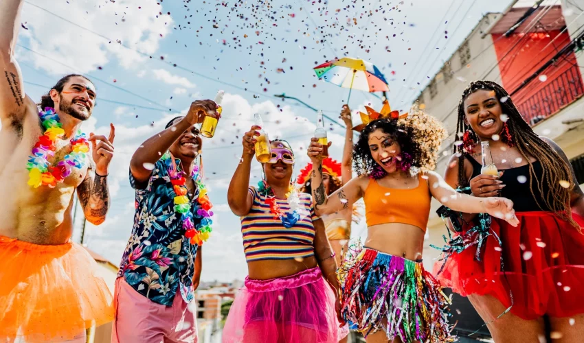 Grupo diversificado de amigos sorrindo, segurando garrafas de bebida e celebrando sob uma chuva de confetes coloridos na rua.