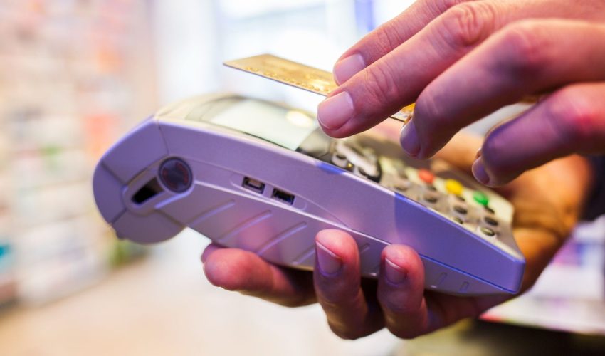 Close-up das mãos de uma pessoa aproximando um cartão de crédito de uma maquininha de pagamento em um ambiente iluminado.