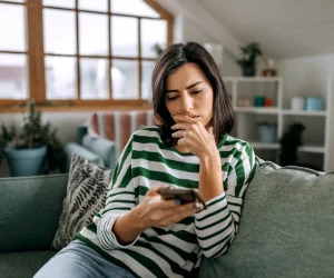 Mulher sentada em um sofá, olhando para a tela do celular com expressão de dúvida e preocupação, com a mão no queixo, analisando uma possível fraude.