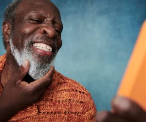 Homem negro idoso sorrindo com satisfação enquanto olha para a tela de um smartphone laranja, demonstrando o uso da tecnologia para cuidar do corpo e da mente e manter a autoestima.