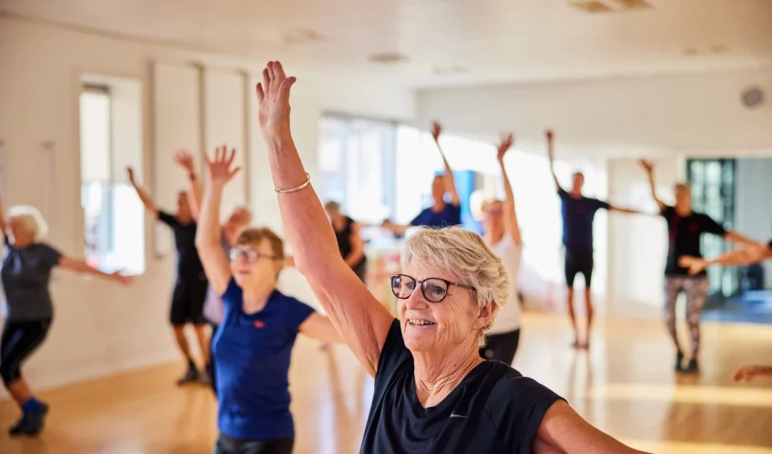 Grupo de idosos em uma sala ampla praticando exercícios de braços levantados, focados na manutenção da mobilidade e no objetivo de cuidar do corpo e da mente.