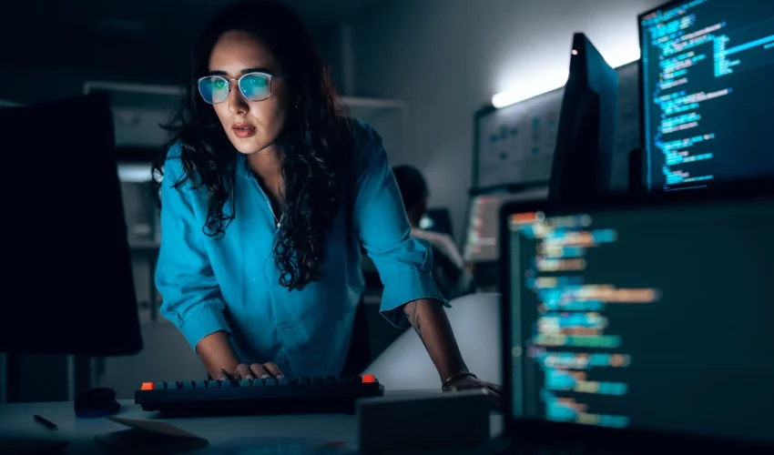 Mulher cientista da computação concentrada diante de monitores com códigos de programação em ambiente noturno de alta tecnologia.