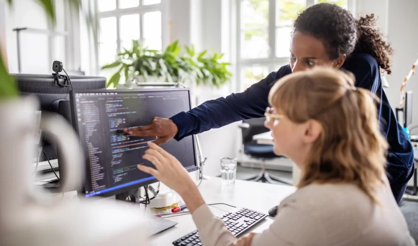 Duas mulheres em um escritório moderno analisando linhas de código complexas em um monitor, colaborando em um projeto de pesquisa tecnológica.