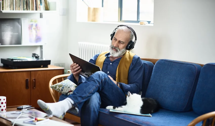 Homem idoso de barba grisalha sentado em um sofá azul, usando fones de ouvido de alta qualidade e segurando um disco, focado em cuidar do corpo e da mente através da musicoterapia.