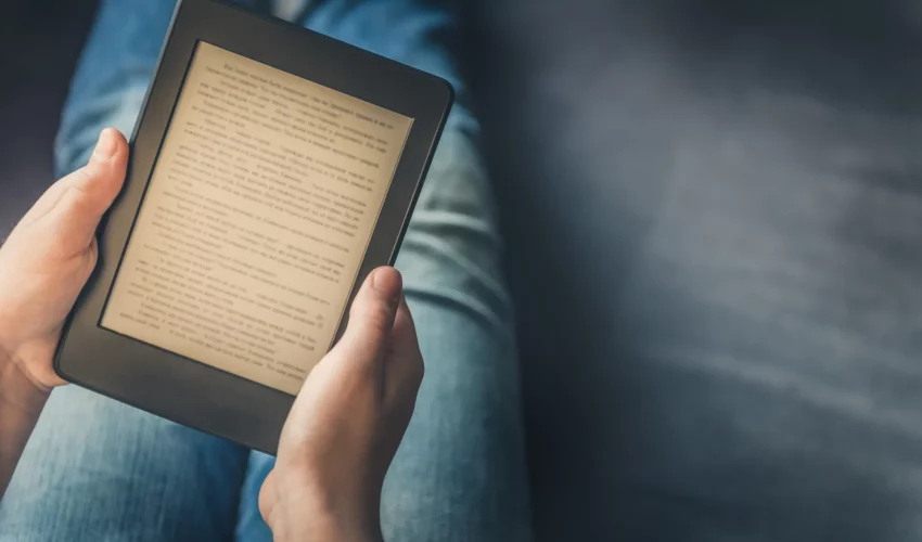 Close-up das mãos de uma pessoa segurando um e-reader (dispositivo de leitura digital) com texto em preto e branco na tela.