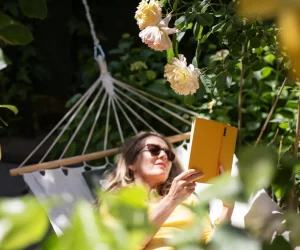 Mulher loira de óculos escuros deitada em uma rede branca, segurando um livro de capa amarela em um jardim ensolarado com flores e limoeiro.