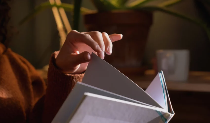 Close-up de uma mão tocando e virando a página de um livro físico de capa dura em um ambiente aconchegante à meia-luz.