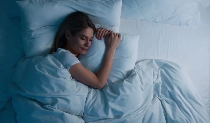 Vista superior de uma mulher loira dormindo tranquilamente em uma cama com lençóis e travesseiros brancos macios, sob uma luz azulada suave.