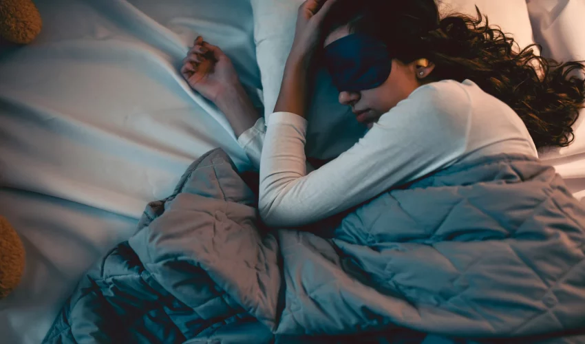 Jovem dormindo de lado usando uma máscara de dormir (tapa-olho) azul escura e protetores auriculares, coberta por um edredom cinza.