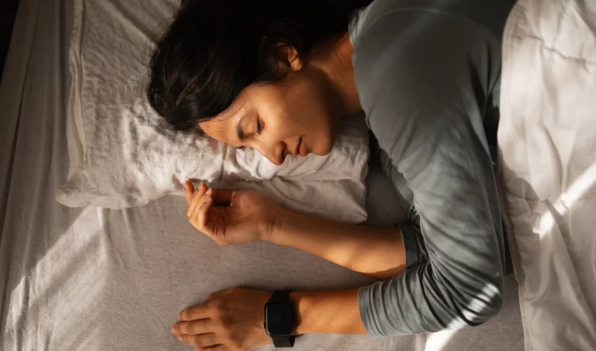 Close em uma mulher dormindo, com foco em seu braço que utiliza um smartwatch de pulseira preta sobre o lençol branco.