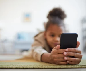 Close-up das mãos de uma criança segurando um celular preto em primeiro plano, com o rosto da criança desfocado ao fundo enquanto ela está deitada em um tapete.