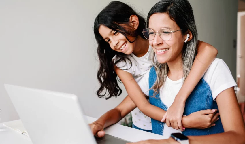 Uma mulher de óculos e fones de ouvido sorri enquanto usa um notebook branco, sendo abraçada por uma criança que também olha para a tela com alegria.