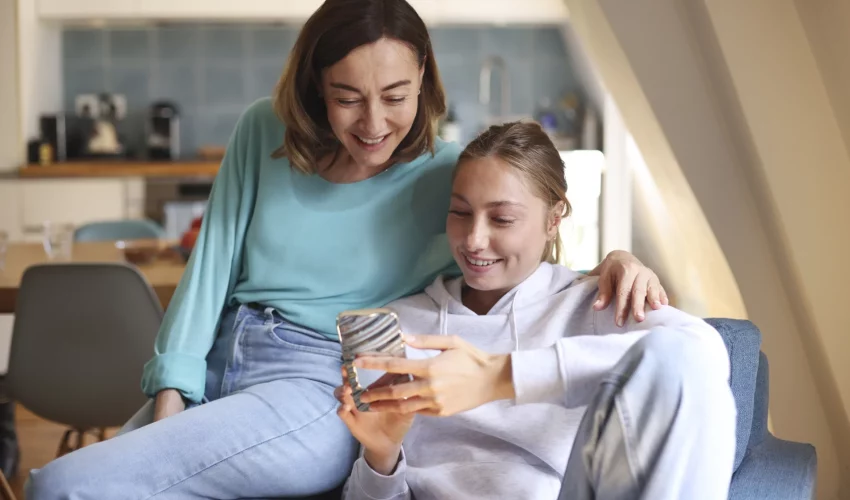 Mulher adulta e uma jovem sentadas no sofá, olhando juntas para a tela de um celular e sorrindo em um ambiente doméstico iluminado.